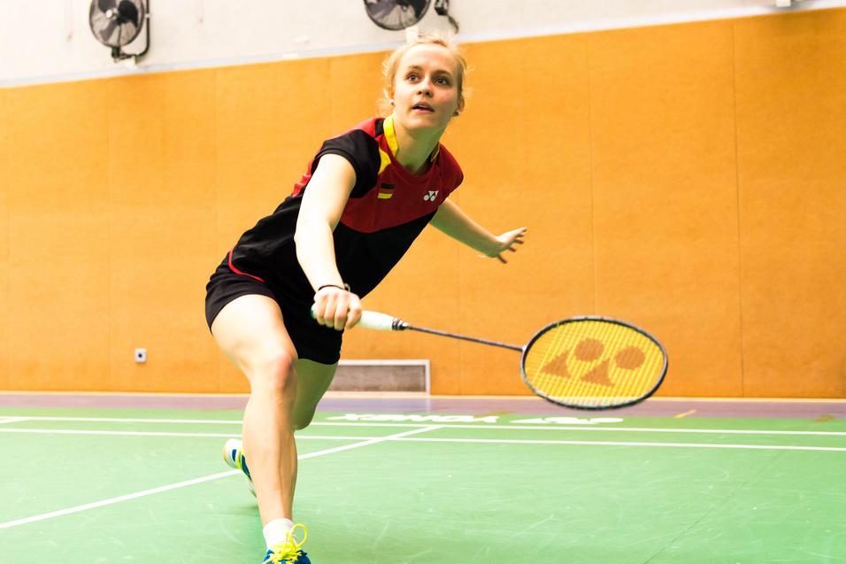 Spielerin schlägt einen Badmintonball auf einer grünen Spielfläche in einer Sporthalle mit orangefarbenen Wänden.