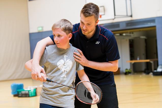Ein Trainer zeigt einem Jungen die richtige Technik beim Badmintonspiel in einer Sporthalle.