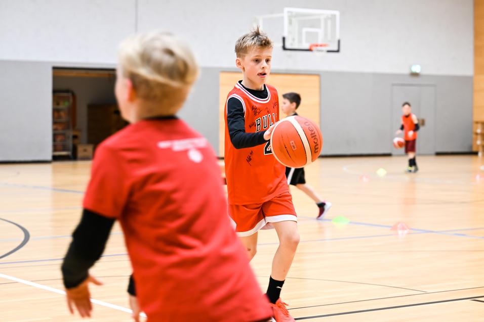 Junge in rotem Basketballtrikot dribbelt einen Ball in einer Sporthalle, während andere Kinder trainieren.