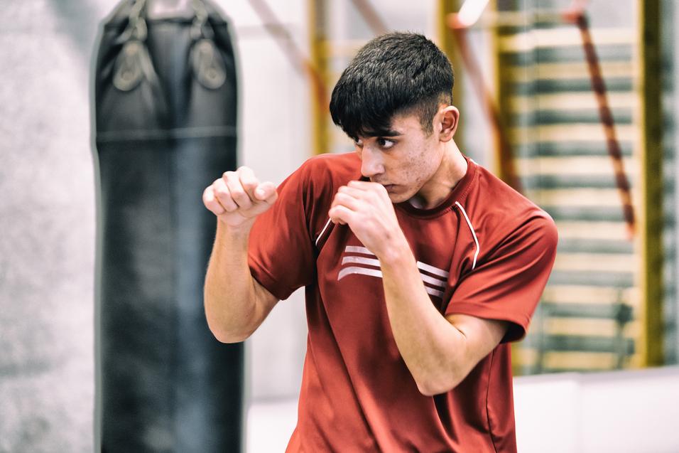 Junger Mann in rotem Trainingsshirt übt Boxbewegungen vor einem Boxsack in einem Trainingsraum.