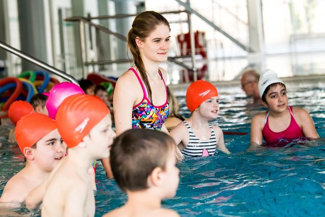 Gruppen von Schwimmkindern mit bunten Badekappen stehen im Wasser, eine Schwimmlehrerin beobachtet sie.